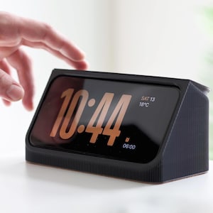 Può includere: Una sveglia digitale nera che mostra l'ora 10:44, sabato 13 e 18°C. L'orologio ha una forma rettangolare con un display inclinato e una texture a coste sui lati. Una mano si avvicina all'orologio.