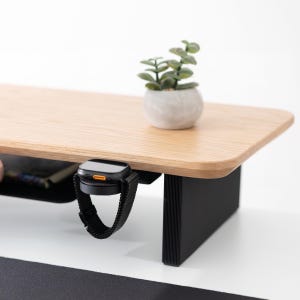 May include: A wooden desk shelf with a black base, featuring a small potted plant and a black smartwatch with a mesh band. The desk shelf has a light wood finish and a rounded edge. The smartwatch has an orange accent.
