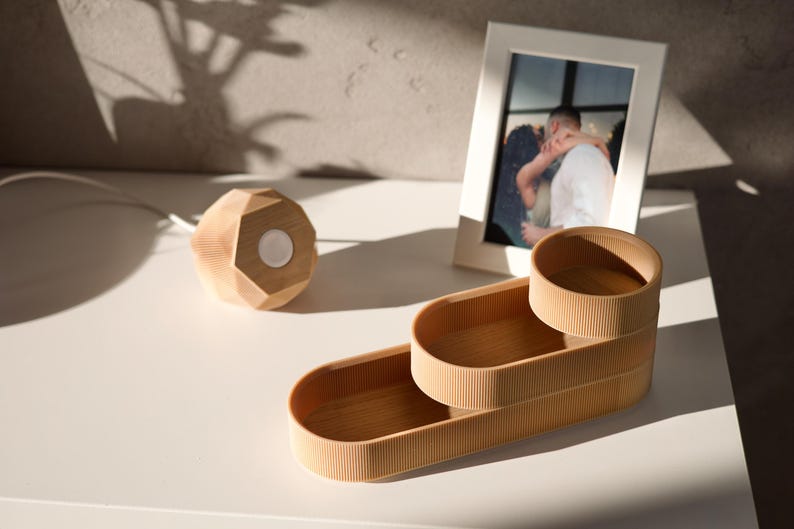 May include: A set of three wooden nesting trays with a rounded rectangular shape. The trays are made of light brown wood and have a ribbed texture. The trays are stacked on top of each other, with the largest tray on the bottom and the smallest tray on top.