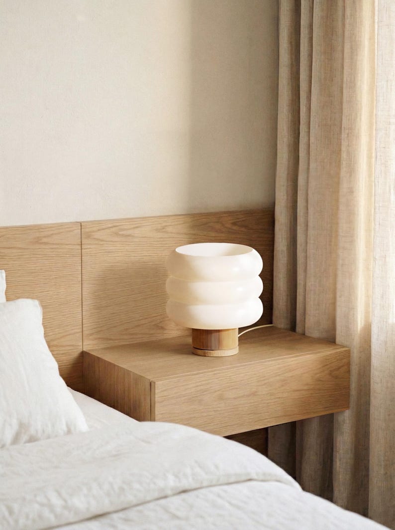 Puede incluir: Una l&aacute;mpara de mesa blanca con anillos apilados y base de madera sobre una mesita de noche de madera. La l&aacute;mpara emite una luz suave y c&aacute;lida. El dormitorio incluye una cama con ropa de cama blanca y una pared y cortina de tonos neutros.
