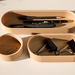 May include: Three wooden trays with rounded edges and a ribbed texture. The top tray holds two black pens and a black marker. The middle tray holds two gold paper clips. The bottom tray holds two black binder clips.