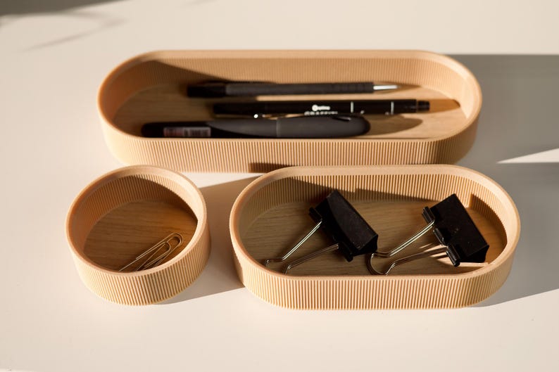 May include: Three wooden trays with rounded edges and a ribbed texture. The top tray holds two black pens and a black marker. The middle tray holds two gold paper clips. The bottom tray holds two black binder clips.