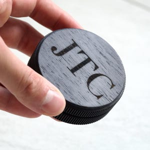Op de afbeelding: Een ronde, donkere houten container met de initialen "JTC" gegraveerd op de bovenkant. De container heeft een geribbelde, zwarte rand en wordt in een hand gehouden. Het hout heeft een grijze tint.