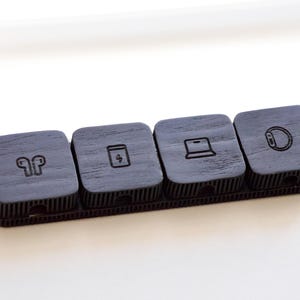 Puede incluir: Un organizador de madera gris oscuro con cuatro compartimentos cuadrados. Cada compartimento tiene un icono diferente: auriculares, un s&iacute;mbolo de carga, un port&aacute;til y un reloj inteligente. Dise&ntilde;ado para peque&ntilde;os accesorios tecnol&oacute;gicos.