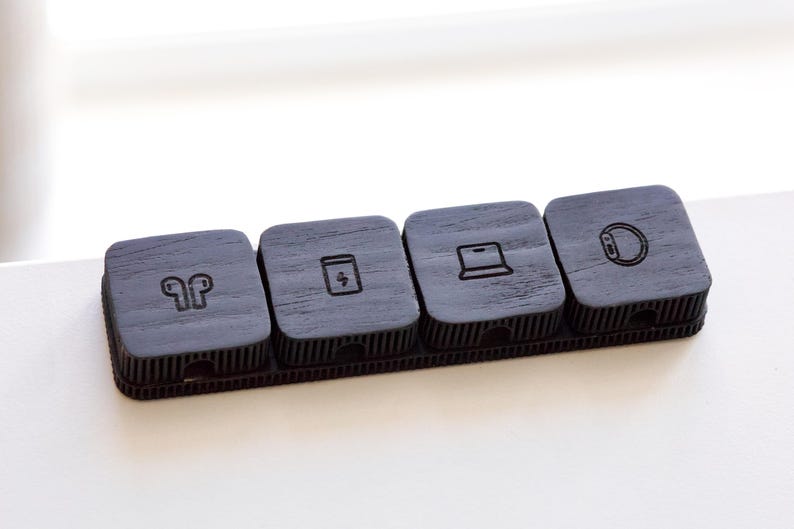 Puede incluir: Un organizador de madera gris oscuro con cuatro compartimentos cuadrados. Cada compartimento tiene un icono diferente: auriculares, un s&iacute;mbolo de carga, un port&aacute;til y un reloj inteligente. Dise&ntilde;ado para peque&ntilde;os accesorios tecnol&oacute;gicos.