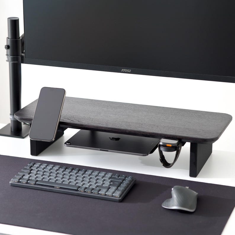 May include: A dark wood monitor stand with a black computer monitor, a smartphone, and a tablet. A wireless keyboard and mouse are on a black desk mat. A smartwatch is attached to the stand.