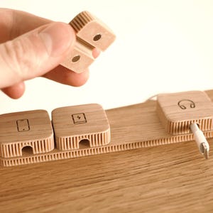 Puede incluir: Organizador de escritorio de madera con tres bloques cuadrados, cada uno con un icono diferente: un tel&eacute;fono, un rayo y unos auriculares. Una mano sostiene un cuarto bloque. El organizador es de madera clara.