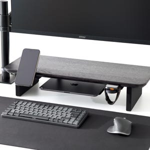 Pode incluir: Um suporte de monitor de madeira preta com uma prateleira embutida, segurando um smartphone, um tablet e um smartwatch. Um teclado e um mouse sem fio estão em um tapete de mesa preto. Um monitor de computador é montado acima.