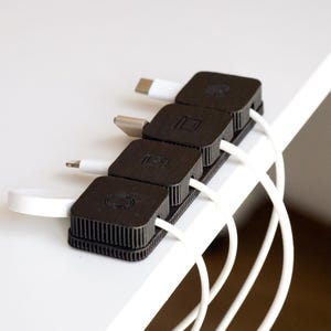 Puede incluir: Un organizador de cables negro con cuatro secciones cuadradas, cada una con un cable blanco. El organizador est&aacute; colocado en el borde de una superficie blanca. Los cables tienen diferentes tipos de conectores.