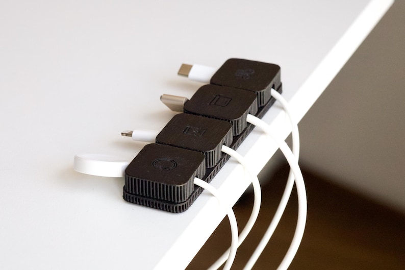 Puede incluir: Un organizador de cables negro con cuatro secciones cuadradas, cada una con un cable blanco. El organizador est&aacute; colocado en el borde de una superficie blanca. Los cables tienen diferentes tipos de conectores.