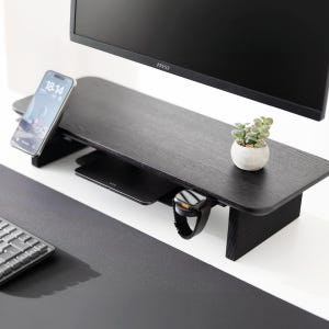 May include: A black wooden monitor stand with a built-in shelf. A smartphone, wireless charger, and smartwatch are on the stand. A small potted plant sits on the right side. A computer monitor is above the stand.