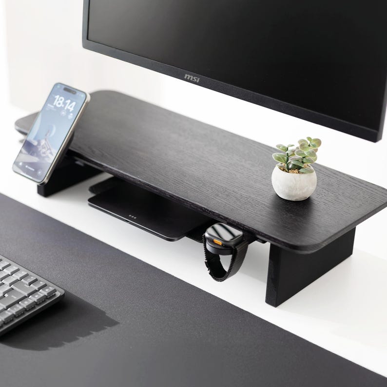 May include: A black wooden monitor stand with a built-in shelf. A smartphone, wireless charger, and smartwatch are on the stand. A small potted plant sits on the right side. A computer monitor is above the stand.