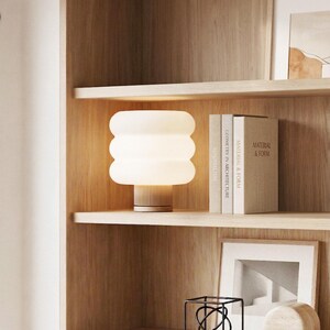 Puede incluir: Una l&aacute;mpara de mesa moderna, blanca, con dise&ntilde;o de globos apilados y base de madera, se encuentra sobre una estanter&iacute;a de madera. La l&aacute;mpara est&aacute; encendida, emitiendo una luz c&aacute;lida. La estanter&iacute;a tambi&eacute;n contiene libros, arte enmarcado y objetos decorativos.