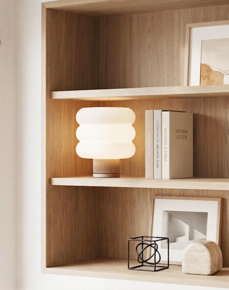 Puede incluir: Una l&aacute;mpara de mesa moderna, blanca, con dise&ntilde;o de globos apilados y base de madera, se encuentra sobre una estanter&iacute;a de madera. La l&aacute;mpara est&aacute; encendida, emitiendo una luz c&aacute;lida. La estanter&iacute;a tambi&eacute;n contiene libros, arte enmarcado y objetos decorativos.