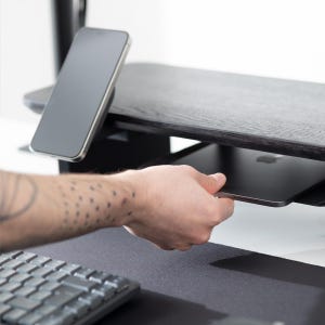 May include: A black wooden desk with a built-in shelf and a smartphone resting on top. A hand is reaching for a tablet on the shelf. A keyboard and mousepad are in the foreground.