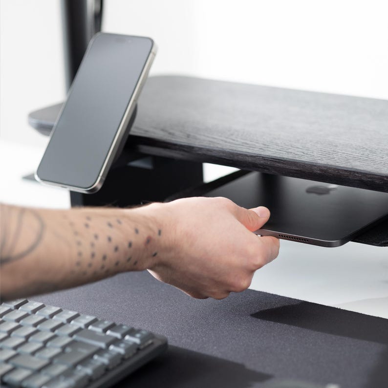 May include: A black wooden desk with a built-in shelf and a smartphone resting on top. A hand is reaching for a tablet on the shelf. A keyboard and mousepad are in the foreground.