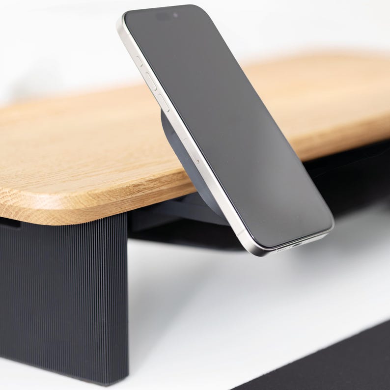 May include: A smartphone with a black screen is mounted on a wooden desk organizer. The desk organizer has a light brown wooden top and black supports. The phone is silver and black.