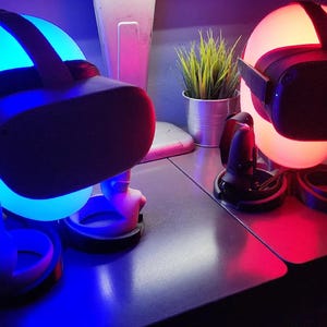 Puede incluir: Dos cascos de realidad virtual, uno azul y otro rojo, se muestran en soportes con controladores. Los cascos están iluminados desde el interior, creando un efecto brillante.