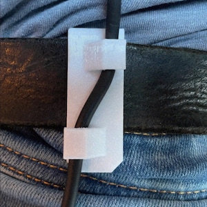Puede incluir: Un clip de cable blanco impreso en 3D que asegura un cable negro a un cinturón negro. El clip está sujeto a unos vaqueros azules. El texto "VIRTUAL REALITY 3D" y una calificación son visibles.