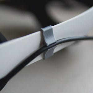 May include: Close-up of a white headset with a black cable secured by a gray clip. The clip is attached to the headset's band, keeping the cable organized. The image highlights the headset's design and cable management.