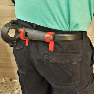 Op de afbeelding: Een zwart en grijs gereedschap met een rode handgreep is bevestigd aan een zwarte riem met een zwarte gesp. Het gereedschap heeft een ronde opening en een handgreep met het woord "PTC" erop gedrukt.