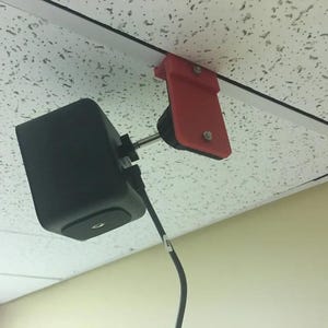 May include: A black square-shaped security camera mounted to a white ceiling with a red plastic bracket. The camera is connected to a black cable.