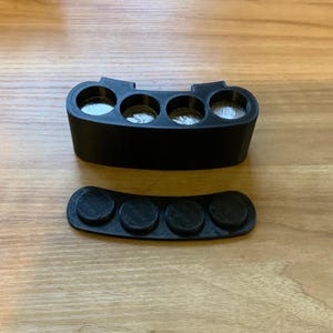 May include: A black 3D-printed coin holder with four circular slots, holding coins. Below it is a matching black tray with four circular indentations. The items are on a light brown wooden surface.