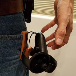 Könnte beinhalten: Ein Holzhalter für einen VR-Controller, der an einer Gürtelschlaufe befestigt ist. Der Halter ist braun und hat einen schwarzen VR-Controller daran befestigt.
