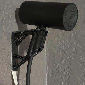Könnte beinhalten: Eine schwarze 3D-gedruckte Wandhalterung für einen VR-Headset-Sensor. Die Halterung hat ein geschwungenes Design, um den Sensor sicher zu halten. Der Sensor ist schwarz und zylindrisch mit einer strukturierten Oberfläche.