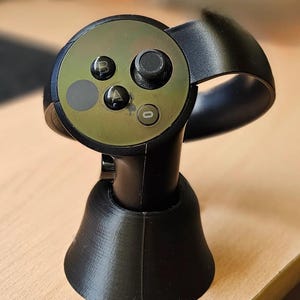 Op de afbeelding: Zwarte VR-headset controller met een cirkelvormige voorkant met knoppen met de labels 'B', 'A' en '0'. De controller heeft een zwarte handgreep en een gebogen steun. Hij staat op een zwarte, uitlopende basis.