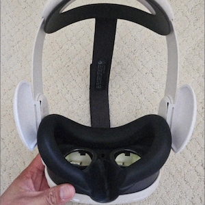 Può includere: Un visore per realtà virtuale bianco e nero con un cuscino facciale nero e auricolari bianchi. Il visore ha un cinturino nero e il testo "VIRTUAL REALITY 3D" è visibile. Il visore è tenuto da una mano.