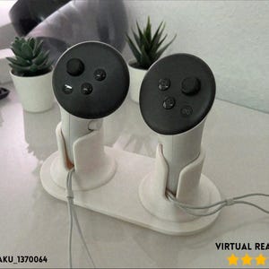 May include: White VR headset controller stand with two cradles. Each cradle holds a controller with a black top featuring buttons. The stand has a rectangular base and is designed to hold VR controllers. The text "VIRTUAL REALITY 3D" is visible.
