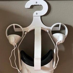 Puede incluir: Un colgador de plástico blanco para auriculares VR con dos correas blancas y una diadema blanca. Los auriculares están colgados del colgador con las lentes mirando hacia la pared.