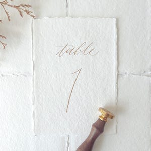 Peut inclure: Une carte de numéro de table en papier blanc avec le mot "table" et le chiffre "1" écrits en calligraphie marron. Un tampon de cire en bois et or est dans le coin inférieur droit. La carte a un bord dentelé.