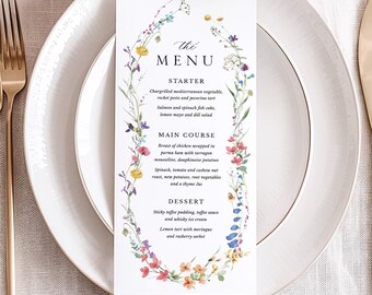 Op de afbeelding: Een formele tafelsetting met een wit bord, gouden bestek en een menukaart. De menukaart heeft een bloemenrand en het woord "MENU" bovenaan. Het menu bevat voorgerechten, hoofdgerechten en dessertopties.