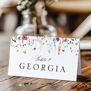 May include: A white table card with a floral watercolour design and the text "Table 4 GEORGIA". The card is folded and stands on a wooden surface. The floral design includes purple, red, yellow, and green.
