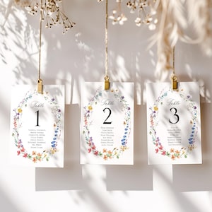 Puede incluir: Tres tarjetas de mesa blancas con diseños de coronas florales y clips dorados. Cada tarjeta muestra un número de mesa y nombres de invitados. Las coronas florales presentan flores y vegetación coloridas, con los números 1, 2 y 3.