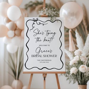 May include: A white sign on a wooden easel with black text that reads "She's Tying the Knot!" and "Welcome to Grace's Bridal Shower." The sign is decorated with a black border and bow. Balloons and flowers are in the background.