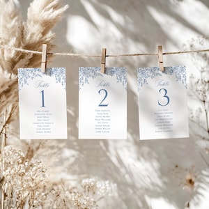 May include: Three white table number cards with blue floral designs and guest names are clipped to a string. The cards are numbered 1, 2, and 3. The cards are hanging in front of a neutral background.