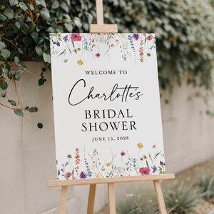 Peut inclure: Un panneau blanc sur un chevalet en bois, avec le texte "Welcome to Charlotte's Bridal Shower" en écriture noire. Le panneau est décoré d'éléments floraux colorés et de la date "June 15, 2028".