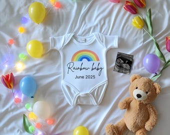 Pode incluir: Um macacão branco com um arco-íris e o texto "Rainbow baby June 2025". O macacão está rodeado de balões coloridos, um urso de peluche e uma corda de luzes.