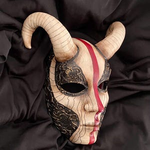 Puede incluir: Una máscara artesanal con base crema, adornada con patrones de filigrana negra y una audaz franja roja en el centro. La máscara presenta cuernos beige curvados y está diseñada con rasgos faciales. Probablemente sea para disfraces o decoración.