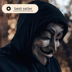 May include: A person wearing a black hooded jacket and a Guy Fawkes mask. The mask is white and has a stylized face with a mustache and a goatee. The text "best seller" is displayed in a white oval.