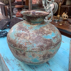 Puede incluir: Un jarrón de metal grande y redondo con una boca ancha y un aspecto envejecido y desgastado. El jarrón tiene una combinación de colores turquesa y marrón, con signos visibles de desgaste y oxidación. Probablemente sea un artículo decorativo antiguo o vintage.