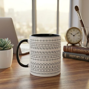 Può includere: Una tazza in ceramica bianca con interno e manico neri, caratterizzata da un motivo geometrico nero ripetitivo di linee orizzontali, quadrati, zig-zag e archi che avvolgono la superficie esterna del recipiente cilindrico.