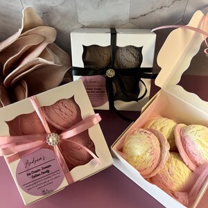 May include: Several boxes of ice cream scoops, with a cotton candy theme. The boxes are white with cut-out windows, tied with ribbons and embellished with decorative brooches. The scoops are pink, yellow, and brown.