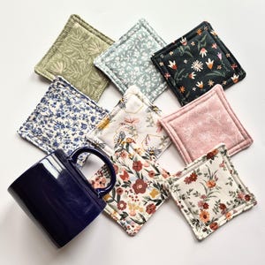 May include: A dark blue ceramic mug positioned next to eight square fabric coasters. The coasters feature various patterns including floral prints in shades of green, blue, pink, and orange, as well as a white coaster with a bee and honeycomb design.