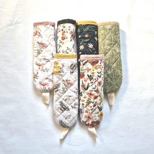 May include: A collection of quilted oven mitts with floral and bee patterns. The mitts feature various colors, including white, pink, yellow, and green, with a quilted design. Each mitt has a loop for hanging.