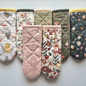 May include: A collection of seven rectangular quilted fabric handle covers for cookware. The covers feature various patterns including bees, honeycombs, floral prints, mushrooms, and botanical designs in colors like white, pink, olive green, and dark gray.