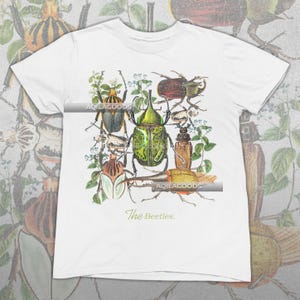 May include: White t-shirt featuring a detailed illustration of various beetles in shades of green, brown, and yellow, with floral accents. The text "The Beetles" is printed in a script font at the bottom. The brand name "AQILAGOODS" is printed on the shirt.